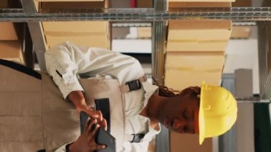 Dikey video: Afro-Amerikalı bir kadın depolama odasında stok yapmak için tablet tutuyor, nakliye ürünlerinden önce malları kontrol etmek için cihaz kullanıyor. Kadın işçi planlama düzeni dağılımı