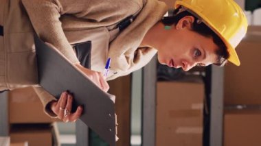 Dikey video: Kadın çalışan depodaki ürünlerin listesini kontrol ediyor, kağıtlarla hisse senedi lojistiği üzerinde çalışıyor ve raflardaki paketleri sayıyor. Depo yöneticisi karton kutuları kontrol ediyor..