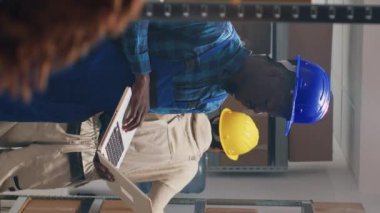 Dikey video: Afro-Amerikalı adam depoda dizüstü bilgisayar kullanıyor, kargoyu karton kutularda kontrol etmek için stok lojistiği üzerinde çalışıyor. Genç yetişkinlerin elinde bilgisayar ve planlama dağıtımı, mallar