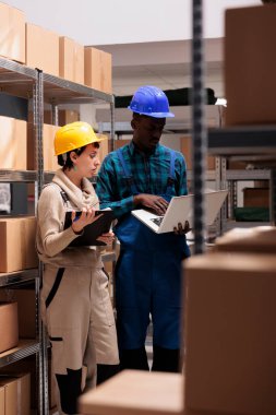 Depo çalışanları dizüstü bilgisayardan müşteri siparişi bilgilerini arıyor ve denetim paketlerini izliyor. Afrika kökenli Amerikalı ve Kafkas depo yöneticileri ellerinde dosya ve bilgisayar yönetimi envanteri tutuyorlar.
