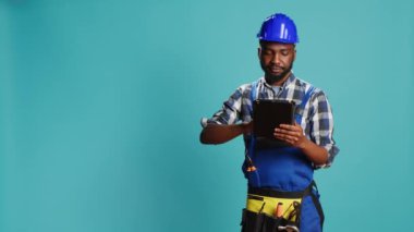 Kamerada dijital tablet kullanan genç tamirci, takımyıldız sözleşmesine hazırlanıyor. Üniformalı erkek marangoz internet sosyal medya sayfasında modern bir cihaz, inşaat sahasında.