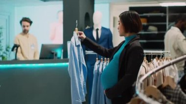 Hamile müşteri askılarda moda kıyafetleri analiz ediyor, gelecekteki anne indirimle almak istiyor. Bebek yumrusu olan genç bir kadın giyim mağazasında şık ürünleri kontrol ediyor, küçük işletmeler..