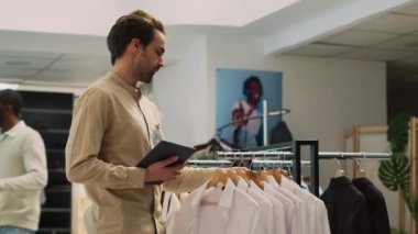 Butik çalışanı mağaza koridorlarının yakınında envanter yapıyor, stokları ve tablet tutan moda kıyafetlerin miktarını kontrol ediyor. Alışveriş merkezindeki mallara bakan genç bir asistan. El kamerasıyla..