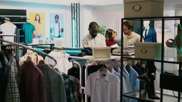 男性の顧客は 流行のファッションコレクションについて従業員に尋ねる 店で新しい商品を検討します ファッショナブルなブティックで服についてショップアシスタントに話をするお店の人 — ストック動画