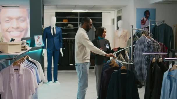 ショッピングセンターで割引商品を検索し モールストアで男性のクライアントにシャツを示すアジアの女性 ブティックショップでディスプレイ上の流行の服を見て女性ショップアシスタント — ストック動画