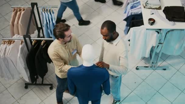 衣料品店でマネキンの正式なスーツを見て店のアシスタントとクライアント モールのブティックで新しい服のための若い男のショッピング 2人の男がファッションコレクションについて話している 三脚ショット — ストック動画