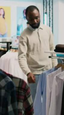Dikey video: Modern adam giyim mağazasında resmi kıyafet alışverişi yapıyor, yeni modern ürünler almak ve gardırobu artırmak istiyor. Moda mağazasını ziyaret eden erkek müşteri, küçük iş konsepti