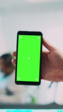 Dikey video: Genç çalışan yeşil ekranlı cep telefonu kullanıyor, akıllı telefondan izole edilmiş telif alanı görüntülerini gösteriyor. Erkek mağaza asistanı butikte boş krom anahtar şablonu tutuyor. Kapat