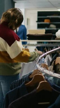 Dikey video: Modern müşteri giyim mağazasından sarkan gömleklere bakıyor, genç kadın gardırobu artırmak için resmi kıyafetler alıyor. Modern moda koleksiyonu satın alan alışveriş merkezi müşterisi.