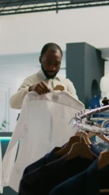 Dikey video: Alışveriş merkezindeki giyim mağazasında asılı duran genç yetişkin inceleme malzemeleri. Moda markalarını satın almak için modaya uygun modern kıyafetlere ya da ürünlere bakan erkek müşteri