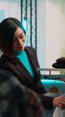 Dikey video: Genç müşteriler şık bir butik dükkanından elbise askıları ve raflardaki resmi giysilere bakıyorlar. Alışveriş merkezinden moda mağaza ürünleri alan kadın, ticari faaliyet.