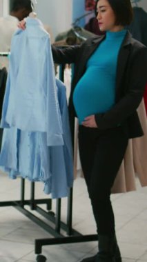 Dikey video: Alışveriş merkezindeki mağazada bebek bezi alışverişi yapan Asyalı kadın askılarda yeni moda markalarına bakıyor. Genç anne hamile. Alışveriş merkezini ziyaret ediyor..