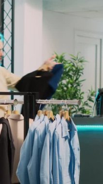 Dikey video: Afro-amerikan müşteri askılarda giysileri inceliyor ve giyim mağazasında malzemeleri kontrol ediyor. Alışveriş merkezinden, perakende mağazasından günlük ya da resmi kıyafet almak isteyen genç bir kadın..