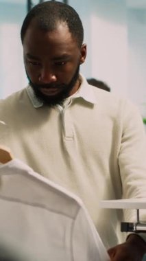 Dikey video: Erkek perakende müşteri raflardan giysiler alıyor, indirimli almak için mağaza ürünlerine bakıyor. Genç yetişkin müşteri olarak alışveriş merkezini ziyaret ediyor, ticari faaliyet