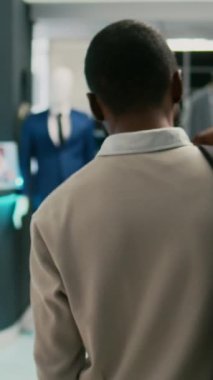 Dikey video: Erkek alışverişçi alışveriş merkezinde resmi giysiler için yeni gömlekler alıyor, gardırobu artırmak için yeni perakende mağaza ürünlerine bakıyor. Delikanlı moda mağazasında moda kıyafetlerini kontrol ediyor..