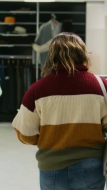 Dikey video: Askılardaki modaya uygun kıyafetleri inceleyen genç kadın, giyim mağazasından resmi veya gündelik giysiler almak istiyor. Alışveriş merkezindeki alışveriş merkezinde yeni malları kontrol eden yetişkin, ticari...