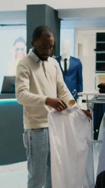Dikey video: Afro-Amerikan müşteri alışveriş merkezindeki mağazada asılı duran resmi gömlek kumaşlarını inceliyor. Erkek müşteri modaya uygun ürünlere bakıyor, mağazadan modern kıyafetler alıyor..