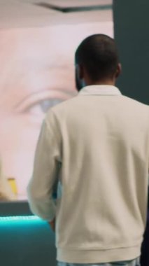 Dikey video: Afrikalı Amerikalı adam yeni modern kıyafetlere para ödüyor, alışveriş merkezindeki posizyon terminalinde işlem yapıyor. Erkek müşteri yazar kasada mağaza asistanıyla konuşuyor, ticari faaliyet