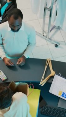 Dikey video: Genç müşteri kredi kartıyla moda kıyafetler alıyor, pos terminalinden alışveriş yapıyor. Alışveriş manyağı bir adam mağazadan kıyafet alıyor, perakende satış elemanıyla konuşuyor. Ticari.