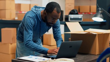 African american worker planning products shipment at warehouse, putting merchandise in boxes for distribution. Employee taking stock goods from storage shelves to ship supplies. Handheld shot.