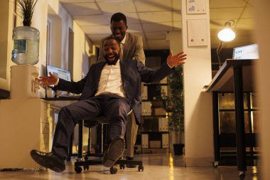 Modern başlangıç ofisinde teneffüste spor yarışlarında masa başı sandalyesine oturan mutlu işçiler. Neşeli Afro-Amerikan yöneticiler eğleniyor, hareketli aktiviteler yapıyorlar.