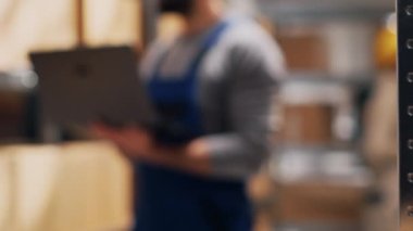 Depo amiri elinde stok lojistik listesiyle dizüstü bilgisayar tutuyor, mal paketlerine bakıyor ve sipariş dağıtımını planlıyor. Kalite kontrol için bilgisayar kullanan bir erkek çalışan. El kamerasıyla..