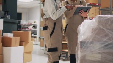 Raflarda ürün organize eden iki kadın, stok lojistik ve planlama dağıtımı için tablete bakıyor. Depo müdürü ve çalışanı karton paketleri sayıyor.