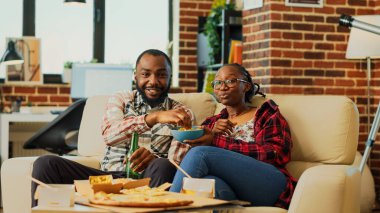 Life partners drinking alcohol and eating snacks while they watch comedy film on television. Young man and woman ordering takeout meal and watching movie on tv, having fun. Tripod shot.