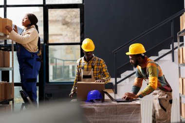 Afro-Amerikan mağaza çalışanları müşteri siparişlerini seçiyor ve dizüstü bilgisayardaki ürün listeleme bilgilerini kontrol ediyor. Depodaki karton kutuyu ararken merdivende duran kadın.