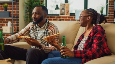 African american people eating slices of pizza in front of tv, binge watching favorite show at home. Relaxed modern couple serving fast food meal and drinking bottles of beer.