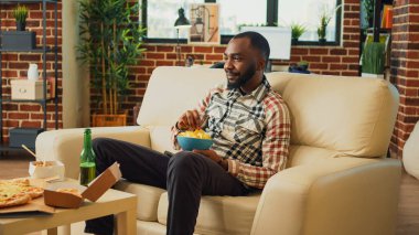 Male adult eating chips from bowl and drinking beer, laughing at comedy film while he has fast food meal on table. Young man serving noodles, hamburger and pizza in living room.