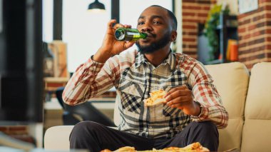 Cheerful young guy drinking beer and eating pizza, watching film on channel program at home. Happy man enjoying dinner with fast food takeout meal and alcohol, watch movie. Handheld shot.