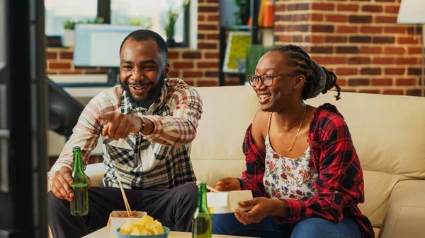 African american couple eating asian food at home, enjoying time together with takeaway meal and beer bottles. Young life partners using chopsticks to eat noodles in living room.