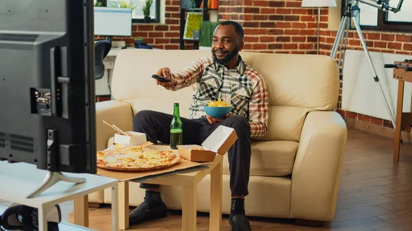 African american adult drinking beer and eating chips, enjoying comedy tv show in living room. Modern man having fun with movie on television, serving takeaway fast food at home.
