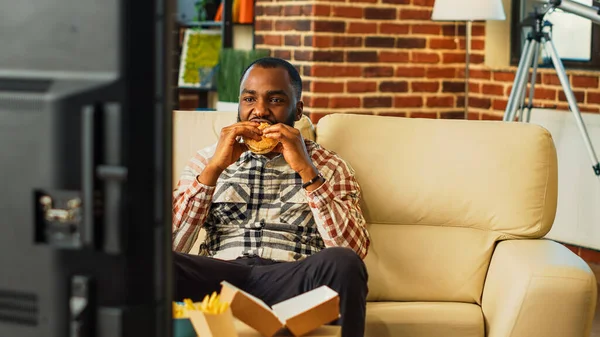 Smiling man eating hamburger with fries and beer, enjoying fast food takeaway meal and watching comedy film on tv. Modern young guy feeling relaxed and happy in front of television.
