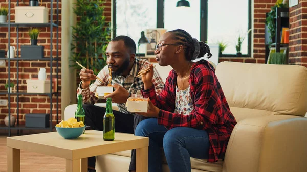 Relaxed couple using chopsticks to eat noodles and watch comedy movie on television. Life partners having fun eating asian food and watching film or tv show in living room, drink beer.