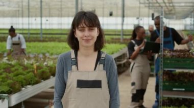 Çevrimiçi siparişler için dizüstü bilgisayar kullanan mühendislerle organik ürün ve sebze çiftliğinde mutlu poz veren kendine güvenen bir kadının portresi. Gülümseyen biyo-tarım işçisi hidroponik mikro yeşillikte duruyor..