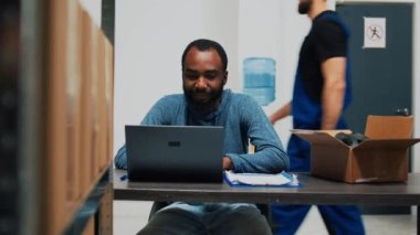 Afro-Amerikalı bir adam mal lojistik ve envanter planlıyordu. Kalite kontrolü için dizüstü bilgisayarla çalışıyordu. Çalışanlar kutularda stok, nakliye ürünleri kullanıyor ve finansal planlama yapıyorlar..