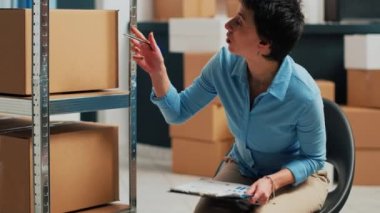 Raflardaki depo mallarını kontrol eden, dosya notlarıyla envanter çıkaran biri. Kadın çalışan stok ürünlerini kontrol etmek ve kalite kontrolü yapmak için kağıtları kullanıyor. El kamerasıyla..