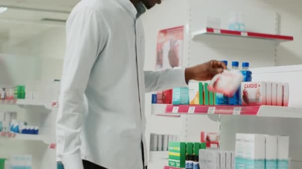 若い男は薬局の棚から薬箱を取って 薬製品を見て治療を購入する クライアントチェックサプリメントパックや医薬品店用品 手持ち撮影 — ストック動画