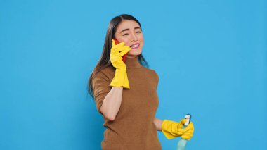 Cheerful housewife wearing protective gloves while holding chemical spray bottle talking at smartphone with remote friend, standing in studio over blue background. Housekeeping and cleaning concept.