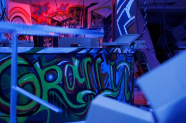 Pembe ve mor neon ışıklarla parıldayan paslı yapısı olan, aydınlık, eski bir bina. Ürkütücü görünüyor. İhmal edilmiş yıkım alanı grafiti spreyi boyası ve floresan parıltısıyla dolu..