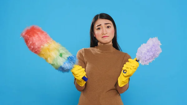 Asian housekeeper looking at professional both feather dusters trying to choose one before start cleaning house. Maid was known for her efficiency and her ability to get the job done quickly