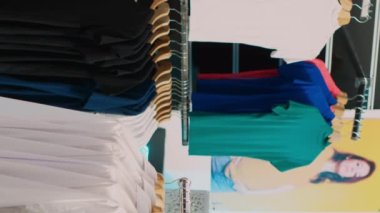 Dikey video: Mağazada elbise askıları ve kumaşları inceleyen kadın müşteri. Genç Alışverişçi, perakende pazarından günlük ya da resmi kıyafet almak istiyor. Üçayak