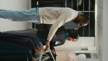 Dikey video: Alışveriş merkezinde indirimli mal arayan Asyalı bir kadın mağazada erkek müşteriye gömlek gösteriyor. Kadın mağaza asistanı butikte moda kıyafetlere bakıyor.