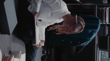 Dikey video: Afro-Amerikan dedektifi yerdeki dava dosyalarına bakıyor, tanık ifadelerini okuyor ve şüpheli kanıtlar buluyor. Sabıka kaydı olan bir kadın müfettiş polis soruşturması yürütüyor..
