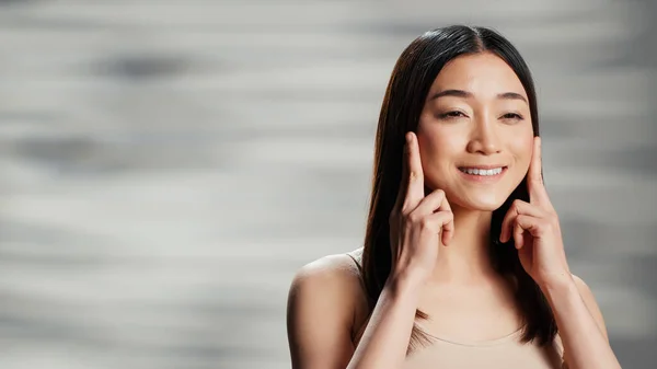 Flawless woman doing beauty routine with facial cream, applying moisturizer on face and advertising skincare cosmetics. Asian female model using sunscreen and promoting self confidence.