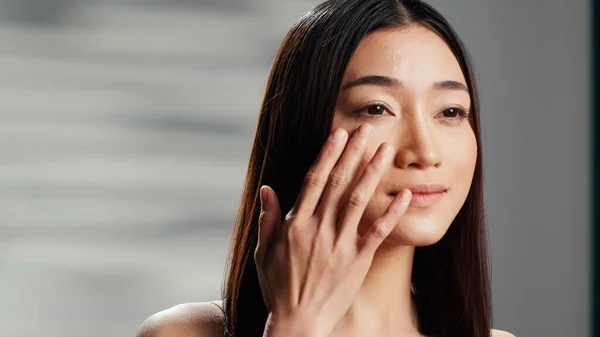Cheerful woman applying anti wrinkles cream on face, promoting dermatology skincare routine cosmetics on camera. Beautiful girl using moisturizer and serum on cheeks, ad campaign.