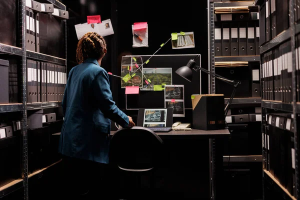 Woman looking at detective board and analyzing crime case evidences. African american woman police forensic inspector investigating proofs and examining clues at night time