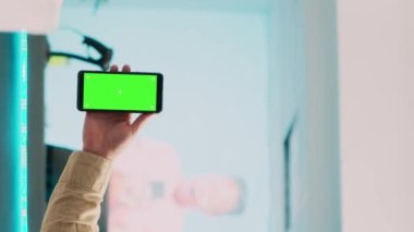 Dikey video: Akıllı telefonu yeşil ekranlı tutan adam cep telefonunda boş telif alanı şablonu gösteriyor. Giyim mağazasında izole edilmiş krom anahtar ekranı kullanan Kafkas pazar asistanı.